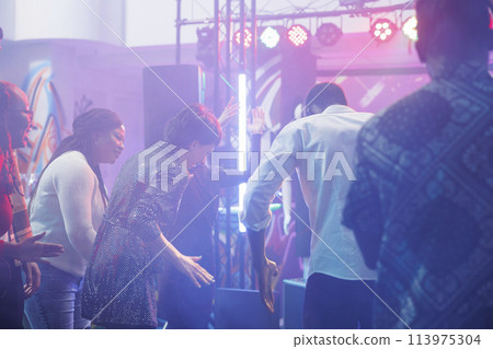 Cheerful clubbers laughing while improvising dance battle at discotheque in nightclub. Carefree man and woman friends showing moves on dancefloor while having fun at club party 113975304