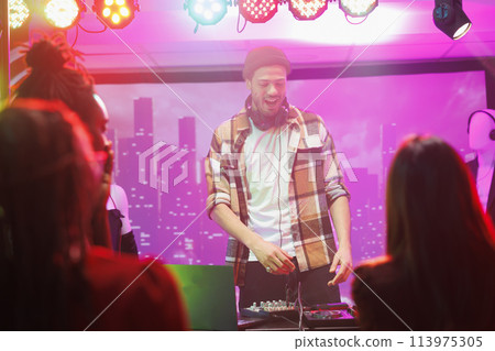 Dj on stage controlling music while crowd partying and dancing in nightclub. Man musician mixing sound on console while performing at live concert in club with spotlights 113975305