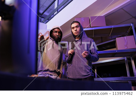 Hackers looking at computer and planning phishing attack. Criminals pointing at screen and discussing ransomware for fraud illegal activity in abandoned warehouse at night 113975462