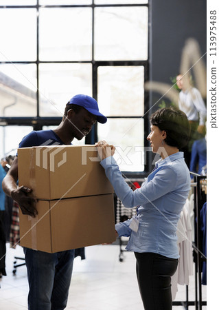 Store manager discussing client orders detalis with african american courier in modern boutique. Man wearing delivery uniform, holding cardboard boxes with stylish clothes in shopping centre 113975488