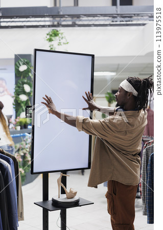 African american man interacting with digital screen offering shoes to customer in shopping center. Clothing store buyer touching white empty display mock up to check stiletto brand options 113975518