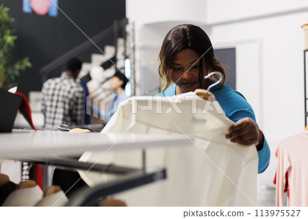 African american customer holding hanger with white shirt, checking items fabric in modern boutique. Shopaholic woman looking at new fashion collection, shopping for casual wear in clothing store African american customer holding hanger with white shirt, checking items fabric in modern boutique. Shopaholic woman looking at new fashion collection, shopping for casual wear in clothing store 113975527