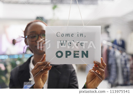 Fashion boutique shop smiling employee hanging we are open sign on store front door. Young african american clothing mall department manager holding signboard with come in message 113975541