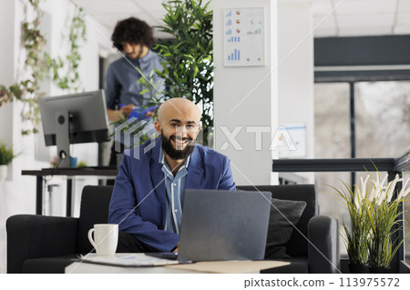 Smiling business entrepreneur working on laptop and looking at camera in start up company office. Successful project manager using portable computer while sitting on couch portrait 113975572