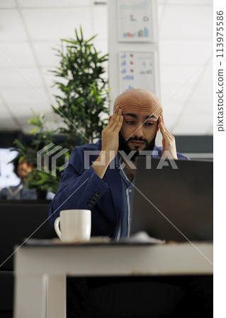 Exhausted arab entrepreneur with migraine writing business plan on laptop in office. Overworked employee having headache while analyzing start up company marketing strategy 113975586