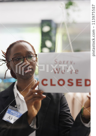 Woman holding closed sign on fashion clothes store front door, informing customers about business hours end. Apparel shop african american assistant hanging sign on widow 113975587