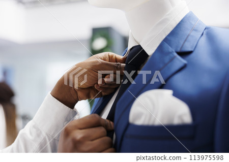 Fashion boutique stylist hands fixing tie and creating formal look on mannequin. African american arms dressing model in suit to display menswear brand in department shopping mall 113975598