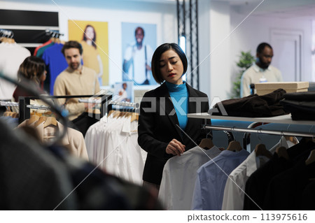 Young asian woman exploring rack with trendy apparel while choosing modern fashionable outfit in shopping mall. Clothing store customer examining formal shirts style in showroom Young asian woman exploring rack with trendy apparel while choosing modern fashionable outfit in shopping mall. Clothing store customer examining formal shirts style in showroom 113975616