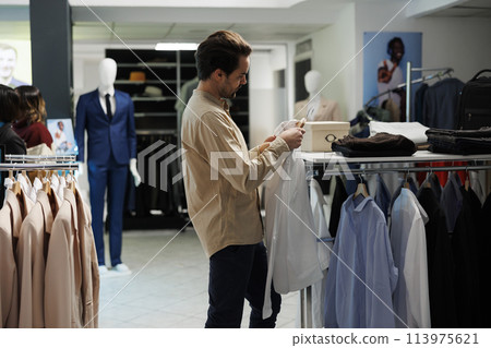 Clothing store customer holding formal shirt on hanger and looking at size label. Young stylish man checking apparel and choosing fashionable garment in shopping mall department Clothing store customer holding formal shirt on hanger and looking at size label. Young stylish man checking apparel and choosing fashionable garment in shopping mall department 113975621