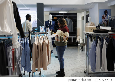 Woman holding shirt on hanger and examining style while shopping in boutique. Young african american buyer searching formal wear and choosing trendy apparel in clothing store 113975623
