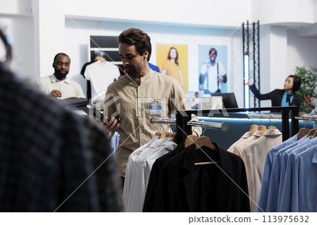 Clothing store manager using digital tablet and organizing merchandise to attract customers. Shopping center employee managing inventory and examining apparel hanging on rack 113975632