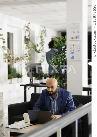 Arab businessman solving marketing strategy problems on laptop while working in start up business office room. Man entrepreneur developing product sales plan on computer in green open space 113975656