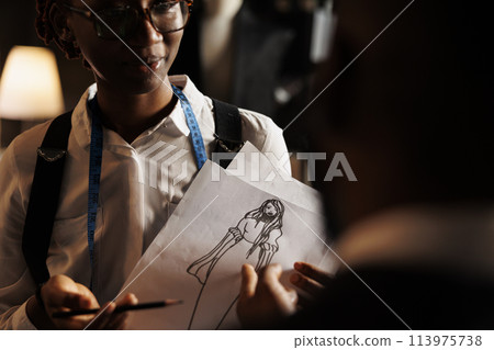 African american couturier showing tailoring studio workspace coworker sketch drawing design she made of haute couture dress. Dressmaker working on comissioned bespoke sartorial outfit African american couturier showing tailoring studio workspace coworker sketch drawing design she made of haute couture dress. Dressmaker working on comissioned bespoke sartorial outfit 113975738