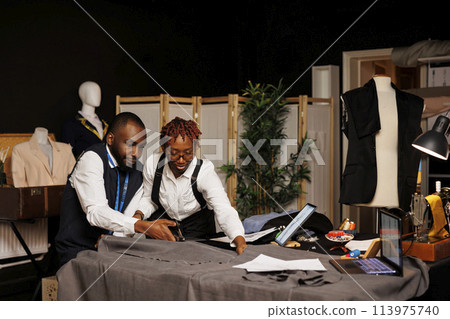 Well dressed couturiers cutting premium fabric in tailoring studio during hand stitched suitmaking process. African american fashion designers preparing textile material for bespoke clothing Well dressed couturiers cutting premium fabric in tailoring studio during hand stitched suitmaking process. African american fashion designers preparing textile material for bespoke clothing 113975740