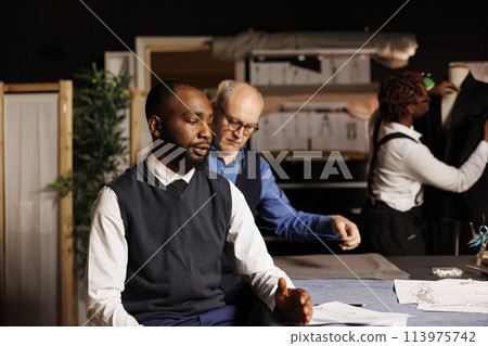 Master tailor taking client measurements while meticulous seamstress apprentice does needlework on suit in background. Gentleman getting custom made attire done by professional couturiers team 113975742