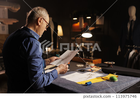 Senior dressmaker conceptualizing outfits for comissioning customer following handdrawn drafts. Elderly tailor looking over sketch drawings, preparing to create bespoken sartorial clothing pieces Senior dressmaker conceptualizing outfits for comissioning customer following handdrawn drafts. Elderly tailor looking over sketch drawings, preparing to create bespoken sartorial clothing pieces 113975746