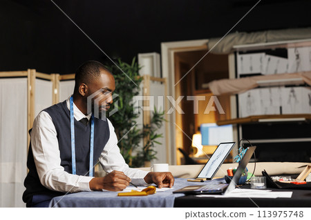 African american dressmaker handdrawing sketch of custom made dress for upcoming exquisite fashion collection. Skilled dressmaker creating bespoken garment piece in atelier shop workspace African american dressmaker handdrawing sketch of custom made dress for upcoming exquisite fashion collection. Skilled dressmaker creating bespoken garment piece in atelier shop workspace 113975748