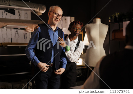 Elderly gentleman shopping for custom made suit happy with outcome in tailoring studio. Senior customer trying on elegant vest helped by african american needleworking seamstress 113975761