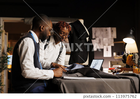 African american tailors working with premium quality fabrics in atelier shop workspace. Happy fashion designers making exquisite garments following sketch designs of upcoming bespoken collection African american tailors working with premium quality fabrics in atelier shop workspace. Happy fashion designers making exquisite garments following sketch designs of upcoming bespoken collection 113975762