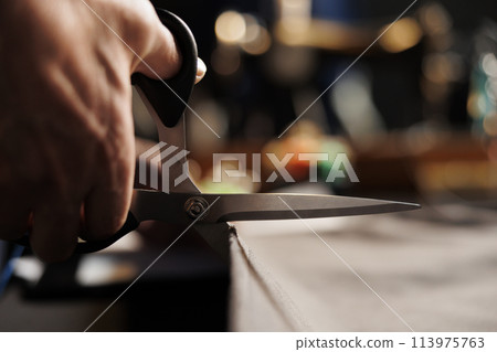Extreme close up of suitmaker using scissors to cut refined textile material in atelier studio, showing expert meticulous craftmanship. Skilled tailor handcrafting bespoke garment piece Extreme close up of suitmaker using scissors to cut refined textile material in atelier studio, showing expert meticulous craftmanship. Skilled tailor handcrafting bespoke garment piece 113975763