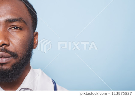 Close-up of African American doctor with a beard looking at the camera. Half-face portrait of black male healthcare specialist wearing white lab coat against isolated blue background. 113975827