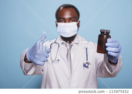 Portrait of medical specialist in a face mask, showing thumbs up while gripping pills bottle. Black doctor with stethoscope and gloves approving prescription painkillers with hand gestures. Portrait of medical specialist in a face mask, showing thumbs up while gripping pills bottle. Black doctor with stethoscope and gloves approving prescription painkillers with hand gestures. 113975832