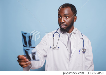 Shocked doctor wearing white lab coat and stethoscope staring at a chest x-ray image that shows alarming test results. Medical professional holding a diagnosis of pulmonary radiography. Shocked doctor wearing white lab coat and stethoscope staring at a chest x-ray image that shows alarming test results. Medical professional holding a diagnosis of pulmonary radiography. 113975850