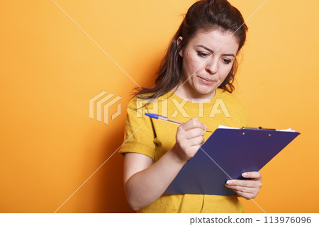 Portrait of female freelancer going through her documents and checking off items on clipboard. Caucasian lady standing in front of isolated background while using a pen to write notes. 113976096