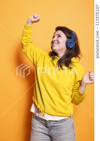 Happy person dancing and listening to music on headphones in studio. Positive woman feeling relaxed and doing dance moves while using headset to enjoy sounds and song over orange background. 113976107