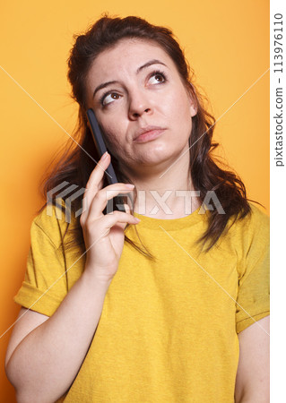 Portrait of female individual having important phone call conversation in front of isolated background. Close-up of woman in thoughts while talking on her mobile device with friends and family. 113976110
