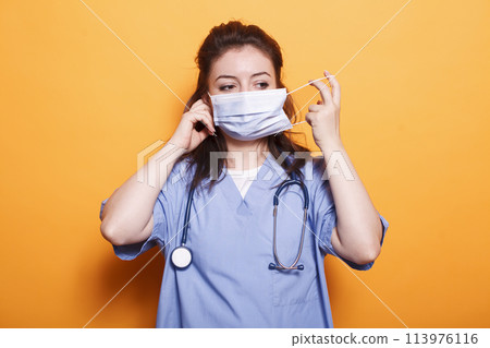 Nurse practitioner putting on face mask for protection in front of camera. Female medical doctor wearing scrubs and stethoscope having safety mask against coronavirus during pandemic. Nurse practitioner putting on face mask for protection in front of camera. Female medical doctor wearing scrubs and stethoscope having safety mask against coronavirus during pandemic. 113976116