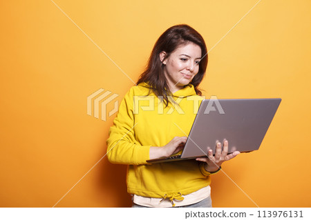 In the studio, young adult looks at a laptop to use technology on camera. Portrait of a modern woman standing over an isolated background, holding a digital device and surfing the internet. In the studio, young adult looks at a laptop to use technology on camera. Portrait of a modern woman standing over an isolated background, holding a digital device and surfing the internet. 113976131