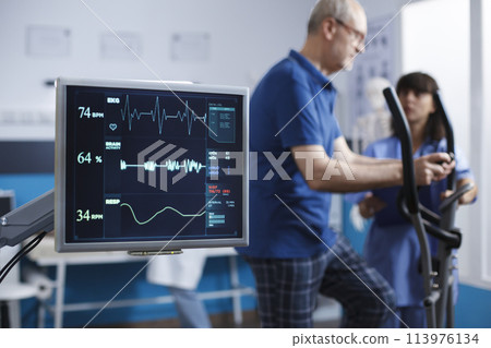 Close-up of heart rate monitor taking a pulse of senior patient during physical rehabilitation activity. Screen showing vital signs of elderly man while he uses stationary bicycle for physiotherapy. 113976134