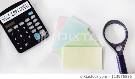 Self-employed word on a calculator, next to a magnifying glass and stickers on a white background 113976886