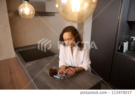 Gorgeous young Latin American brunette using mobile phone for online communication. Happy Arab woman browsing social networks on smartphone and drinking tea in the kitchen at home. Free space for ads Gorgeous young Latin American brunette using mobile phone for online communication. Happy Arab woman browsing social networks on smartphone and drinking tea in the kitchen at home. Free space for ads 113978037