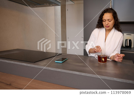 Beautiful curly brunette in white bathrobe, standing at kitchen counter and adding sugar to hot drink. Charming young woman stirring sugar in a cup of tea while having a breakfast in the home kitchen. 113978069