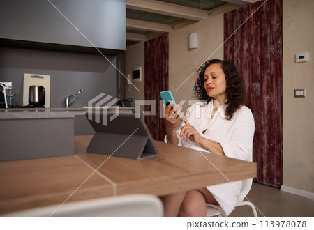 Attractive young woman in white bathrobe, checking news on her smartphone, sitting at table with digital tablet in the morning at home Attractive young woman in white bathrobe, checking news on her smartphone, sitting at table with digital tablet in the morning at home 113978078