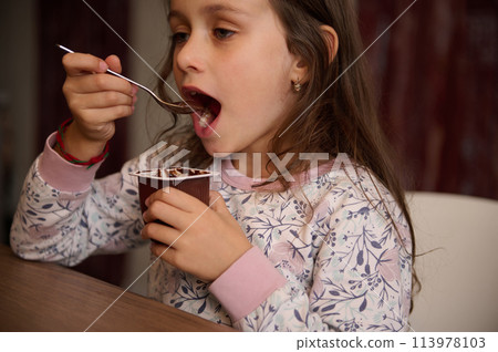Caucasian little kid girl snacking with tasty yoghurt at home. Food and drink consumerism. Authentic portrait of a lovely child 6 years old, enjoying the taste of chocolate vegan yogurt. Caucasian little kid girl snacking with tasty yoghurt at home. Food and drink consumerism. Authentic portrait of a lovely child 6 years old, enjoying the taste of chocolate vegan yogurt. 113978103