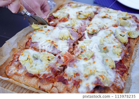 Man cutting homemade artichoke and ham pizza with a pizza cutter 113978235
