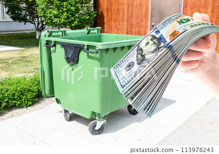 Big green plastic garbage container and stack of american dollars in the hand 113978243