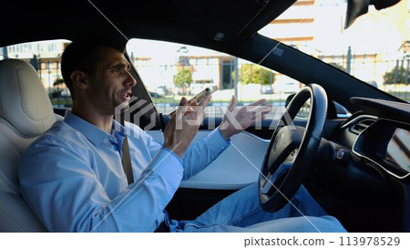 Male businessperson have serious telephone conversation while riding an autonomous self driving electric car. Nervous businessman talks by smartphone during riding on electrical auto with autopilot 113978529