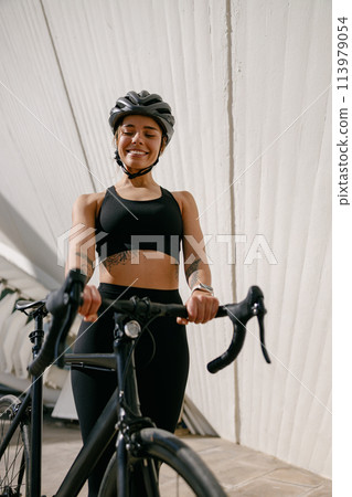 Smiling woman cyclist in protective gear standing with her bike while training outdoors Smiling woman cyclist in protective gear standing with her bike while training outdoors 113979054