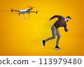 Young man in casual clothes running from camera drone that chases him on amber background. 113979480