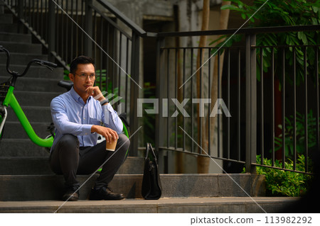 Thoughtful office worker guy looking away while sitting on stairs near office building 113982292