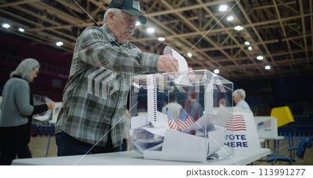 Elderly voter puts bulletin into box and starts look at camera Elderly voter puts bulletin into box and starts look at camera 113991277