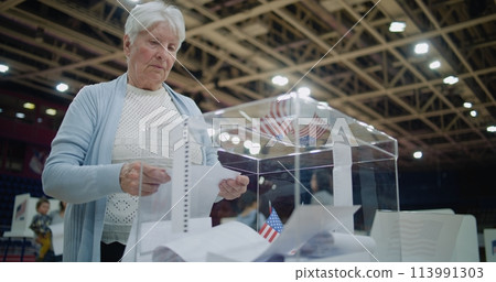 Elderly female voter puts bulletin into box, looks at camera Elderly female voter puts bulletin into box, looks at camera 113991303