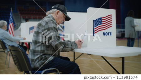 Elderly male voter sits at voting booth in polling station 113991574