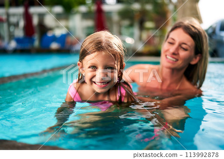 Smiling girl in pink swimsuit learns swimming in pool, mother supports. Child first swim lesson, joyful family moments. Summer day, poolside bonding, teaching kids, healthy lifestyle, water safety. 113992878