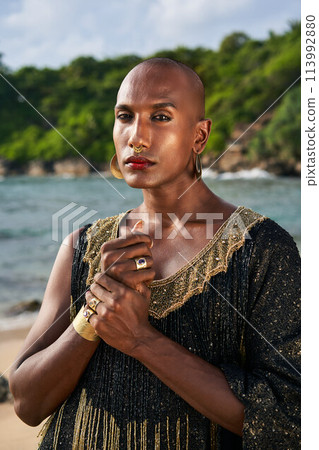 Gorgeous gay black man in luxury gown, jewelry poses on scenic ocean beach . Non-binary ethnic fashion model wears long posh dress golden rings, earrings, bracelets, looks at camera. Gorgeous gay black man in luxury gown, jewelry poses on scenic ocean beach . Non-binary ethnic fashion model wears long posh dress golden rings, earrings, bracelets, looks at camera. 113992880