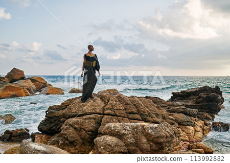 Non-binary black godlike person poses gracefully standing on rocks in ocean. Trans ethnic fashion model in posh dress and jewellery on rocky beach by storm . Lgbtq. Pride month Non-binary black godlike person poses gracefully standing on rocks in ocean. Trans ethnic fashion model in posh dress and jewellery on rocky beach by storm . Lgbtq. Pride month 113992882